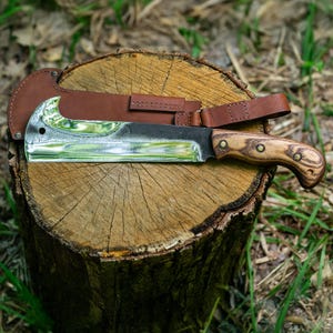 May include: A large, polished steel machete with a wooden handle and a brown leather sheath. The blade has a curved edge and a decorative pattern. The handle is secured with rivets. The machete rests on a tree stump.
