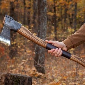 Könnte beinhalten: Eine Person hält eine Axt mit einem Holzstiel und einem schwarzen Ledergriff. Die Axt hat einen dunkelgrauen Metallkopf und einen leicht gebogenen Stiel. Die Axt wird vor einem verschwommenen Hintergrund aus Bäumen und Blättern gehalten.