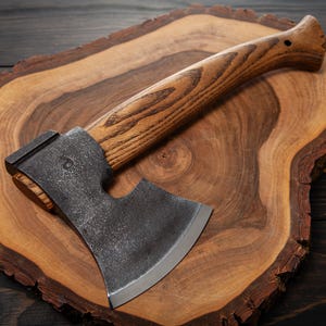 Può includere: Un'ascia nera e argentata con manico in legno si trova su un tagliere di legno. L'ascia ha una lama curva e un piccolo foro nel manico.
