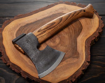 May include: A black and silver axe with a wooden handle sits on a wooden chopping board. The axe has a curved blade and a small hole in the handle.