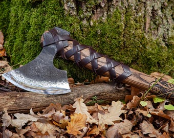 May include: A hand-forged axe with a leather wrapped handle and a Viking knot design. The axe is resting on a log with autumn leaves scattered around it.