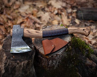 Può includere: Un'ascia forgiata a mano con manico in legno e fodero in pelle. L'ascia è appoggiata su un ceppo d'albero con muschio.