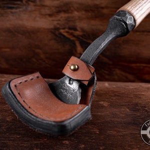 May include: A small, hand-forged axe with a leather sheath. The axe has a wooden handle and a black metal head. The sheath is brown leather with a black stitching detail. The axe is resting on a wooden surface.
