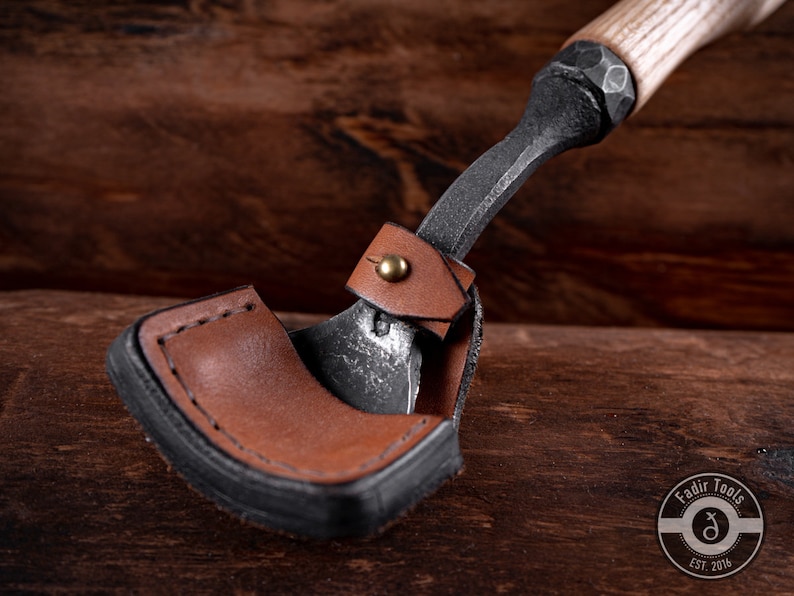 May include: A small, hand-forged axe with a leather sheath. The axe has a wooden handle and a black metal head. The sheath is brown leather with a black stitching detail. The axe is resting on a wooden surface.