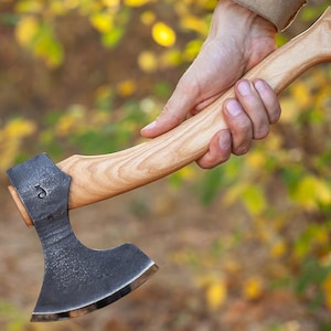May include: A hand holds a black axe with a wooden handle. The axe has a curved blade and a small metal ring on the handle. The background is blurred and out of focus.