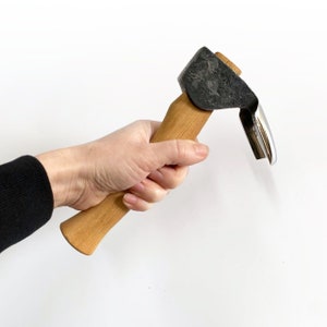 Peut inclure: Un outil de sculpture avec un manche en bois, une tête en métal noir et une lame argentée. L'outil est tenu dans une main.