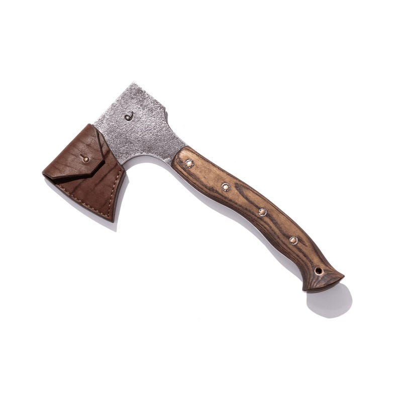 May include: A small, rustic axe with a wooden handle and a metal head. The handle is a dark brown color with silver rivets. A brown leather sheath protects the axe head. The axe is designed for outdoor use.