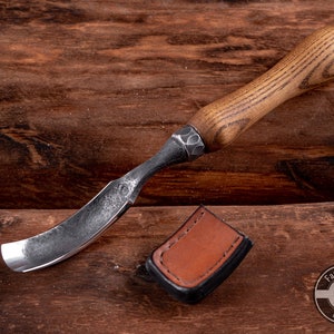 May include: A curved wood carving tool with a dark brown wooden handle and a black metal blade. The tool is lying on a wooden surface with a brown leather sheath.