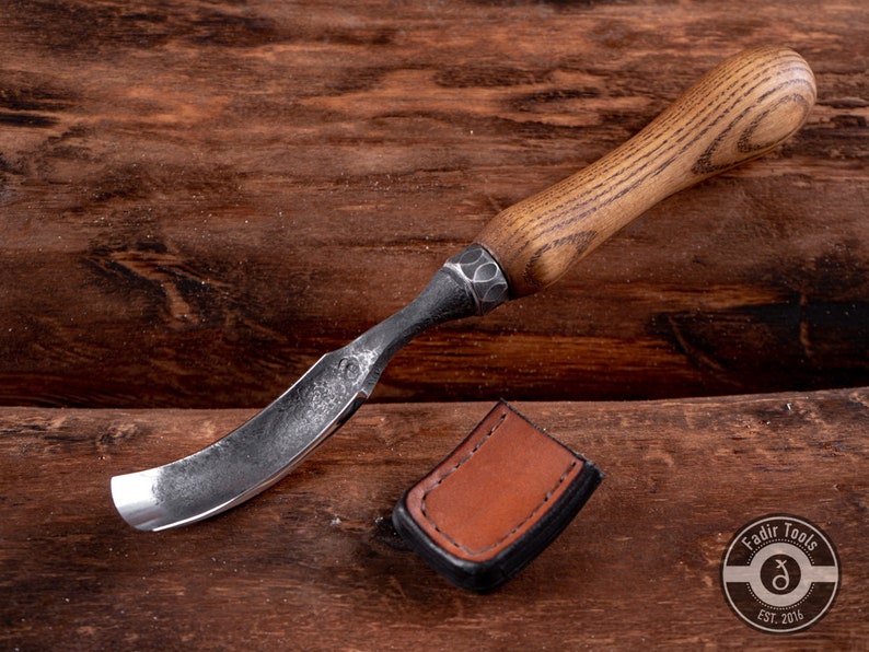 May include: A curved wood carving tool with a dark brown wooden handle and a black metal blade. The tool is lying on a wooden surface with a brown leather sheath.