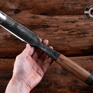 May include: A long, curved, metal chisel with a wooden handle. The chisel has a dark gray finish and a rounded blade. The handle is made of light brown wood with a dark brown metal cap. The chisel is held in a hand against a wooden background.