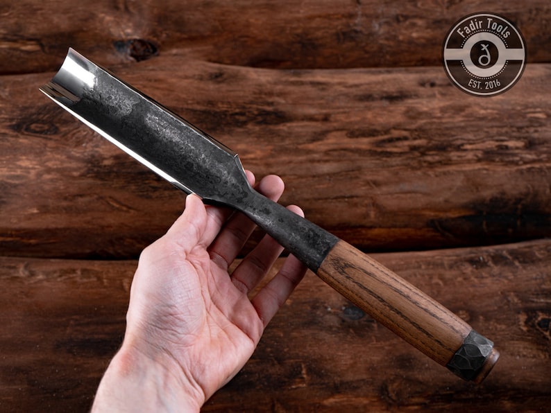May include: A long, curved, metal chisel with a wooden handle. The chisel has a dark gray finish and a rounded blade. The handle is made of light brown wood with a dark brown metal cap. The chisel is held in a hand against a wooden background.