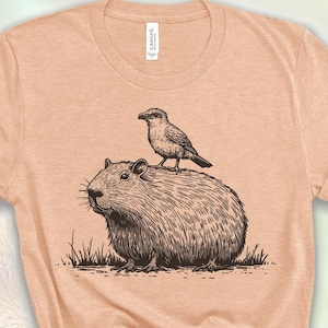 May include: A light peach colored t-shirt with a black and white illustration of a capybara with a bird perched on its back.