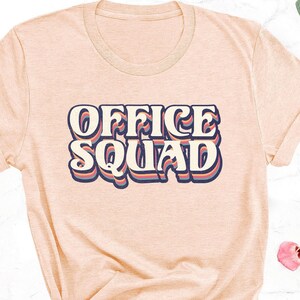 May include: Peach-colored t-shirt with the text "OFFICE SQUAD" in a retro font. The text is white with a blue and red outline. The shirt is styled with a knot at the bottom and surrounded by floral arrangements.