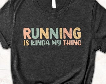May include: Dark gray t-shirt with the text "RUNNING is kinda my thing" in pastel colors. The shirt is knotted at the bottom. The shirt is laying on a white surface with books and greenery in the background.