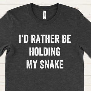 May include: Dark gray t-shirt with the text "I'D RATHER BE HOLDING MY SNAKE" in white block letters. The shirt has short sleeves and a crew neck. The background is a light wood grain.