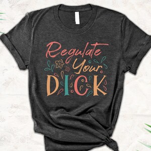 Pode incluir: T-shirt cinzenta escura com a frase "Regulate Your Dick" em letras coloridas e estilizadas. O design inclui detalhes florais e de folhas em tons de coral, verde-azulado e laranja. A t-shirt está atada na parte inferior.