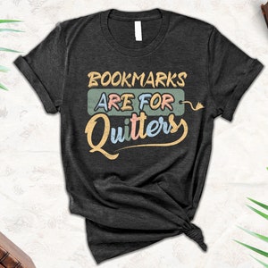 May include: A dark gray t-shirt with the text "Bookmarks are for Quitters" in a colorful retro font.