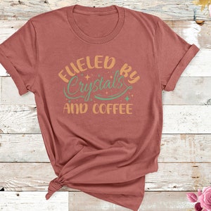 Op de afbeelding: Een roestkleurig T-shirt met de tekst "Fueled by Crystals and Coffee" in een vintage-geïnspireerd lettertype. De tekst is in tinten oranje, groen en wit.