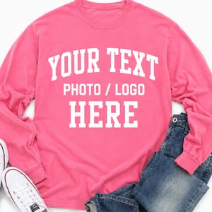 Op de afbeelding: Een felroze sweatshirt met lange mouwen en ronde hals, voorzien van witte blokletters met de tekst YOUR TEXT PHOTO / LOGO HERE, gecombineerd met een blauwe spijkerbroek en witte canvas sneakers met witte veters en rubberen zolen.