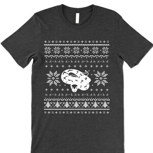 May include: A dark gray t-shirt with a white Christmas sweater design featuring a snake. The design includes snowflakes, reindeer, and Christmas trees.