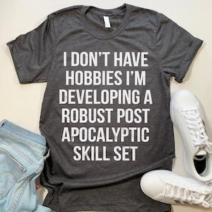 May include: A dark gray t-shirt with white text that reads "I don't have hobbies I'm developing a robust post apocalyptic skill set".