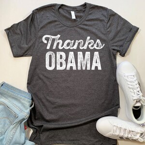 Op de afbeelding: Een donkergrijs T-shirt met de tekst "Thanks Obama" in wit gedrukt op de voorkant. De tekst heeft een verweerde, vintage look.