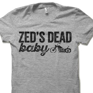 May include: A heather gray t-shirt with the text "ZED'S DEAD baby" in distressed black lettering. A motorcycle graphic is printed to the right of the text. The shirt has a crew neck and short sleeves.