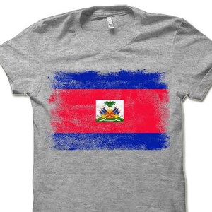 May include: A gray t-shirt with a distressed, faded design of the Haitian flag. The flag features the colors blue, red, and white, with the coat of arms in the center.