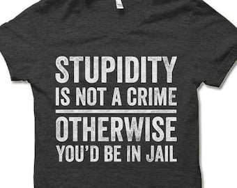 May include: A dark gray t-shirt with a white graphic print that reads "Stupidity is not a crime otherwise you'd be in jail".