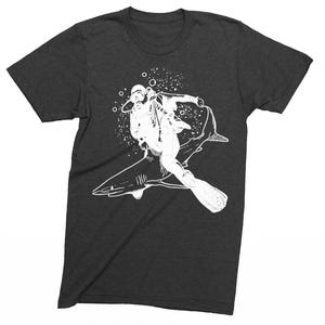 May include: Dark gray t-shirt featuring a white graphic of a scuba diver riding a shark. The diver is wearing a diving helmet, mask, and fins. Bubbles surround the diver and shark, creating a dynamic underwater scene.