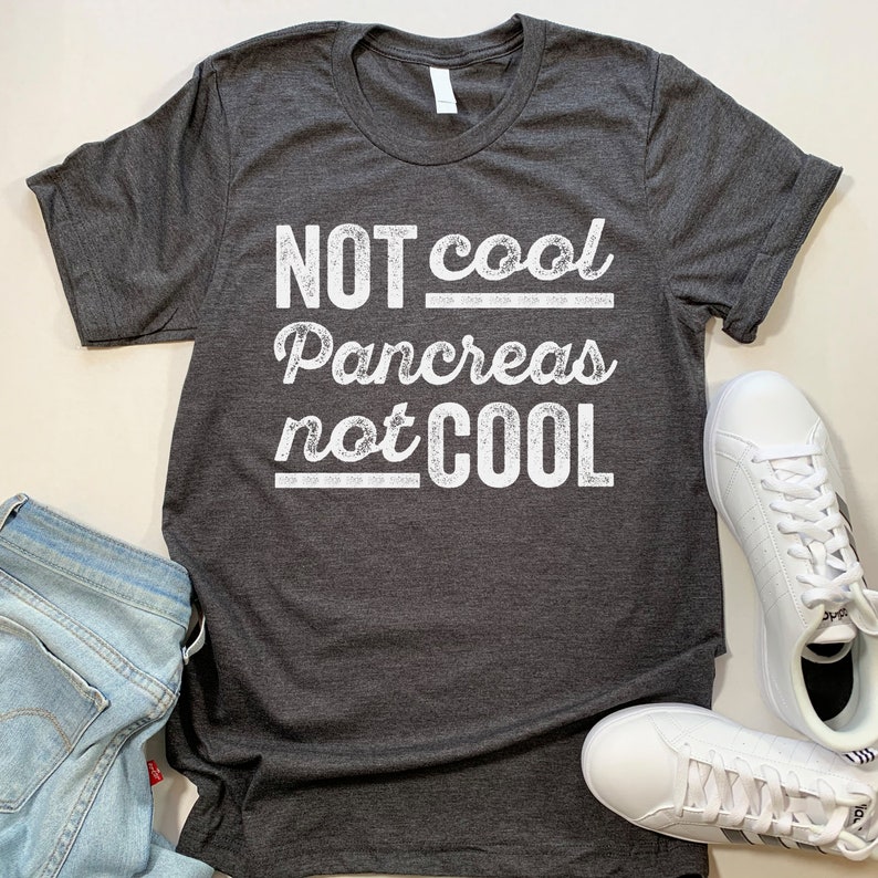 K&ouml;nnte beinhalten: Ein dunkelgraues T-Shirt mit einem wei&szlig;en, abgenutzten Aufdruck, der "NOT cool Pancreas not COOL" lautet.
