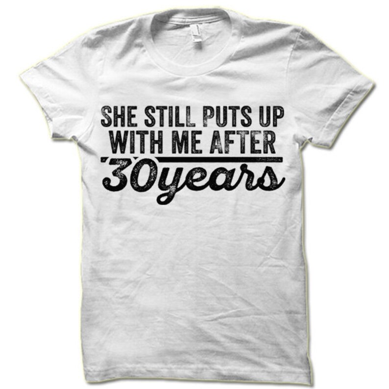 May include: White t-shirt with black text that reads "She still puts up with me after 30 years".