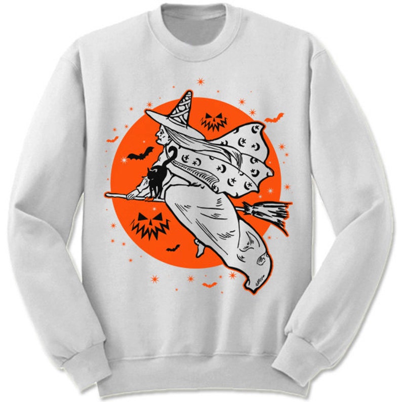 May include: A white sweatshirt with a black and white illustration of a witch flying on a broomstick in front of an orange moon. The witch is wearing a pointed hat and has a black cat on her shoulder. The moon has stars and bats around it.