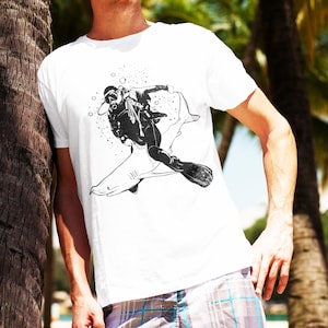 May include: White t-shirt featuring a black and white graphic of a scuba diver riding a shark. The diver is wearing a diving mask and flippers. The design includes bubbles and is set against a tropical background.