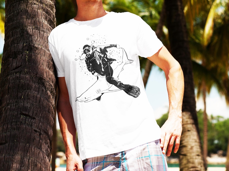 May include: White t-shirt featuring a black and white graphic of a scuba diver riding a shark. The diver is wearing a diving mask and flippers. The design includes bubbles and is set against a tropical background.