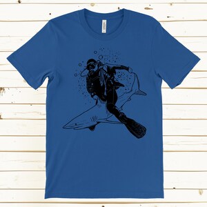 May include: Blue t-shirt featuring a black graphic of a scuba diver riding a shark. The diver is wearing a diving suit and gear. The shark is gray with white accents. Bubbles are in the background.