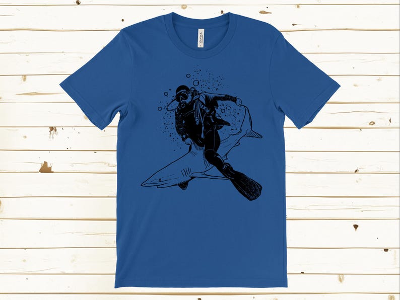 May include: Blue t-shirt featuring a black graphic of a scuba diver riding a shark. The diver is wearing a diving suit and gear. The shark is gray with white accents. Bubbles are in the background.