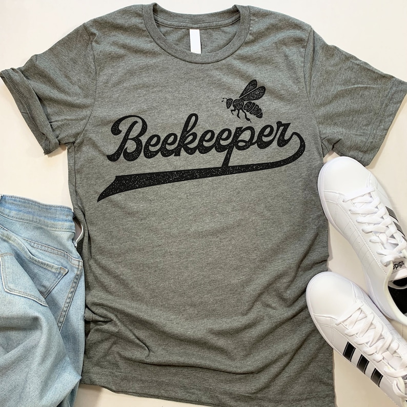 Puede incluir: Una camiseta gris jaspeada con un gr&aacute;fico de abeja negra y el texto "Beekeeper" en una fuente de b&eacute;isbol vintage.
