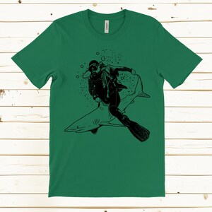 May include: Green t-shirt featuring a black graphic of a scuba diver riding a shark. The diver is wearing a diving suit, mask, and fins. Bubbles surround the diver and shark. The shirt is made of a soft material.