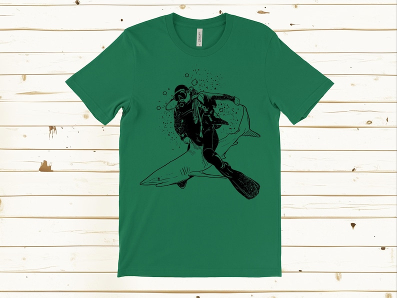 May include: Green t-shirt featuring a black graphic of a scuba diver riding a shark. The diver is wearing a diving suit, mask, and fins. Bubbles surround the diver and shark. The shirt is made of a soft material.
