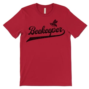 Puede incluir: Camiseta roja de manga corta con un gr&aacute;fico de abeja negra y el texto "Beekeeper" en una fuente de estilo vintage de camiseta de b&eacute;isbol.