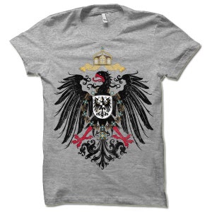 May include: A grey t-shirt with a black and red eagle design. The eagle is holding a shield with a black eagle on it. The eagle is surrounded by a crown and a floral design.