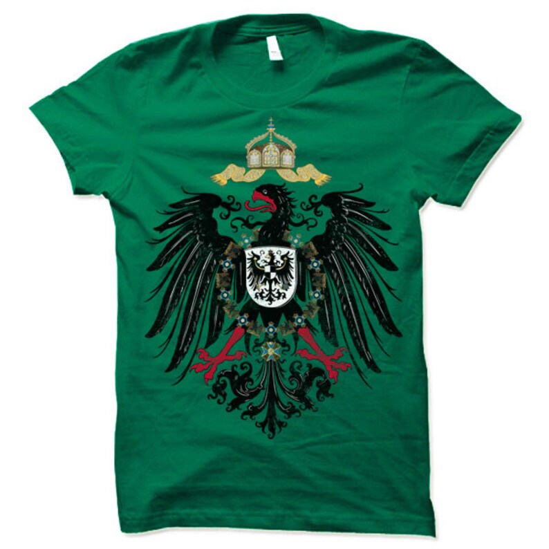 May include: A green t-shirt with a black and gold eagle design. The eagle is holding a shield with a black cross on a white background. The eagle is surrounded by a wreath of flowers and leaves.