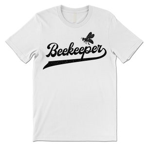 Puede incluir: Camiseta blanca con un gr&aacute;fico negro de una abeja y la palabra "Beekeeper" en una fuente de b&eacute;isbol vintage.