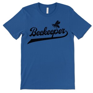 Puede incluir: Una camiseta azul real con un gr&aacute;fico de abeja negra y el texto "Beekeeper" en una fuente de b&eacute;isbol vintage.