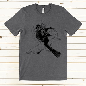 May include: Dark gray t-shirt featuring a black graphic of a scuba diver riding a shark. The diver is wearing a diving suit, mask, and fins. Bubbles and splatters surround the diver and shark. The shirt is made of a soft material.