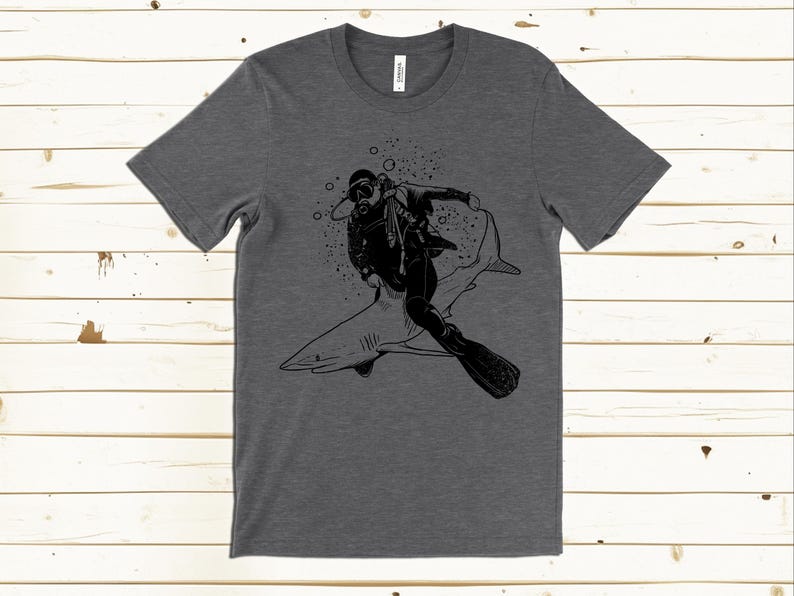 May include: Dark gray t-shirt featuring a black graphic of a scuba diver riding a shark. The diver is wearing a diving suit, mask, and fins. Bubbles and splatters surround the diver and shark. The shirt is made of a soft material.