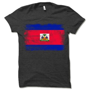 May include: A dark grey t-shirt with a distressed, faded design of the Haitian flag. The flag features the colors red, blue, and white, with a coat of arms in the center.