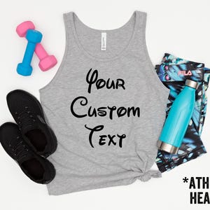 May include: A gray athletic heather tank top with the text "Your Custom Text" in black script. The image also includes black sneakers, pink and blue dumbbells, a teal water bottle, and patterned leggings.