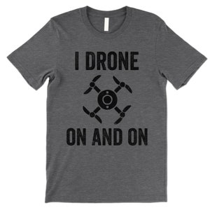 Op de afbeelding: Een donkergrijs T-shirt met de tekst "I DRONE ON AND ON" in zwart. Een zwart drone-icoon is gecentreerd tussen de twee regels tekst.